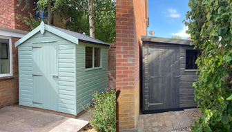 A sage green apex roof shed on a patio to the left, and a dark grey pent roof shed tucked beside a house beneath a tree on the right